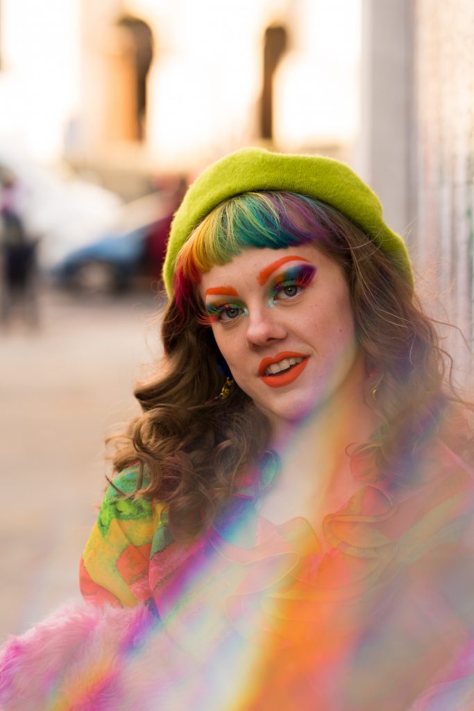 Person with rainbow hair, rainbow eye makeup, red eyebrows, and a rainbow top.