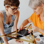 Woman in orange shirt standing beside woman in yellow shirt and holding piece of black marble